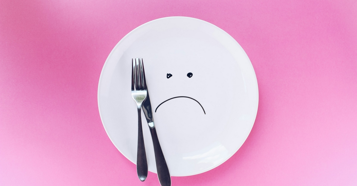 A dinner plate featuring a sad face drawn on with black pen. A knife and fork are on top of the plate.