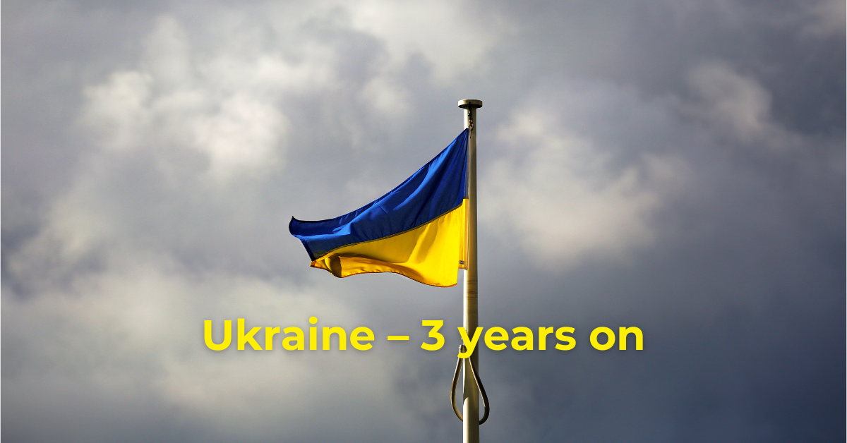 Ukraine – Three years on. Text superimposed on a Ukrainian national flag on a flag pole against a cloudy sky.