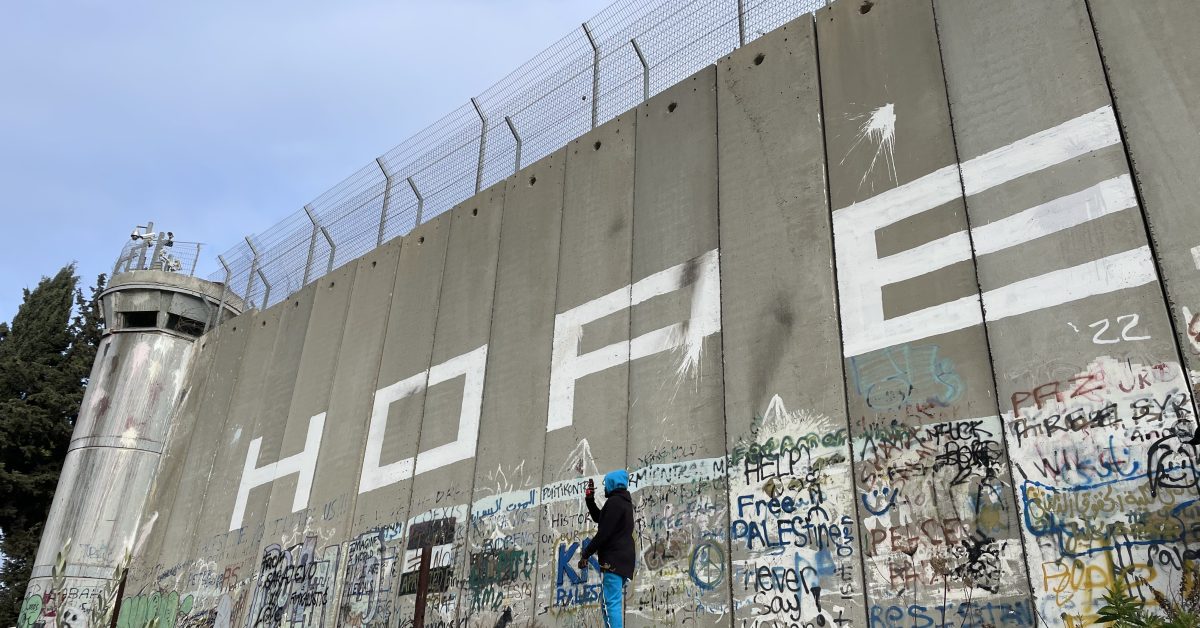 A large wall with military lookout post separating Palestinian and Israeli areas in Bethlehem. It is topped by wire and covered in graffiti. A hooded photographer snaps the graffiti. The word 'Hope' is written in very large letters halfway up the wall.