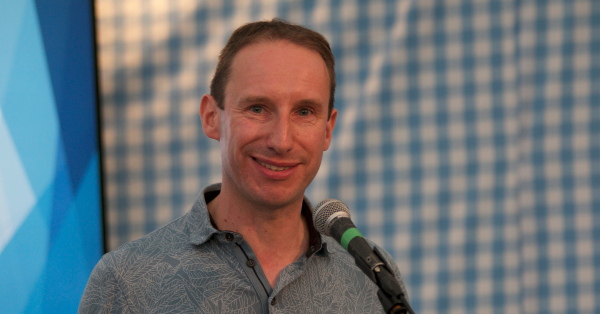 Simeon Mitchell in front of a microphone. In the background is a gingham checked backcloth inside a large marquee.