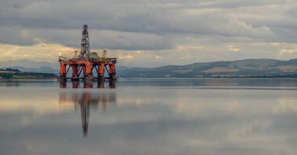 Offshore oil drilling platform standing in calm water beneath a cloudy sky, with its orange support structure and tall derrick reflected on the glassy surface. Low hills and distant shoreline stretch across the background, while muted sunlight breaks through the clouds.
