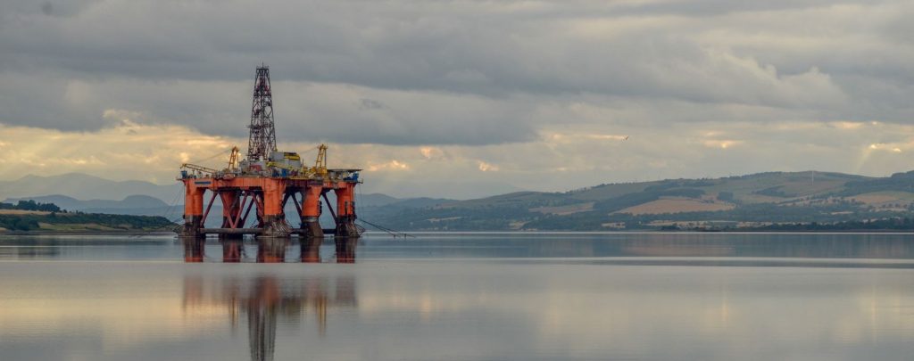 Offshore oil drilling platform standing in calm water beneath a cloudy sky, with its orange support structure and tall derrick reflected on the glassy surface. Low hills and distant shoreline stretch across the background, while muted sunlight breaks through the clouds.