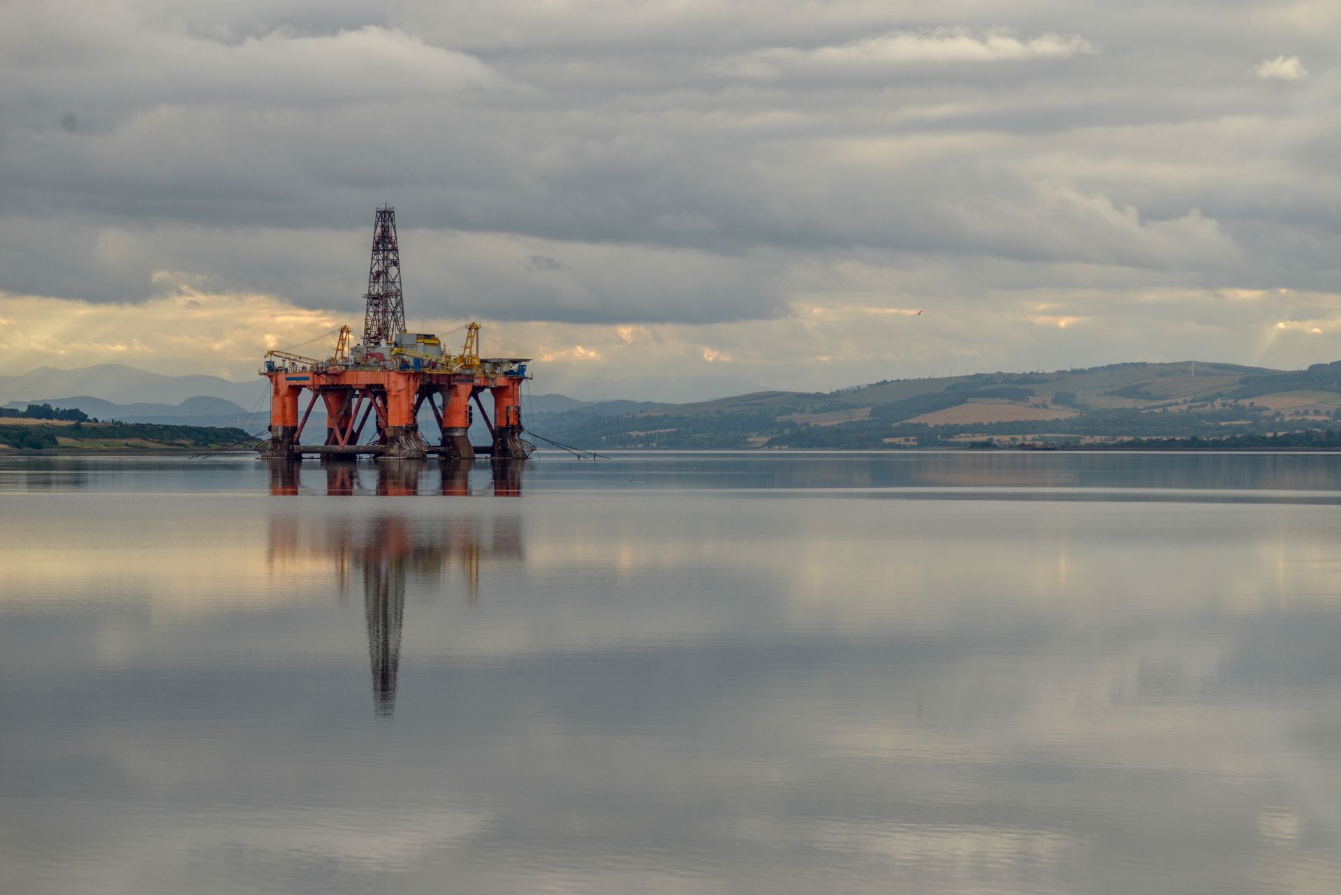Offshore oil drilling platform standing in calm water beneath a cloudy sky, with its orange support structure and tall derrick reflected on the glassy surface. Low hills and distant shoreline stretch across the background, while muted sunlight breaks through the clouds.